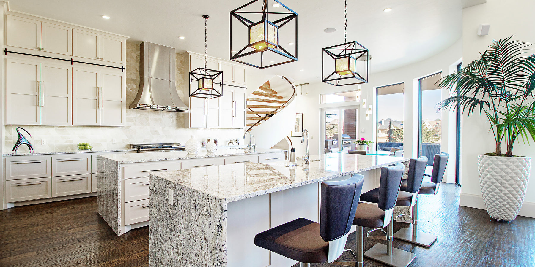 Modern kitchen with a large marble island, sleek white cabinetry, and stylish pendant lighting. A spiral staircase leads to the upper level, with floor-to-ceiling windows providing natural light and outdoor views.